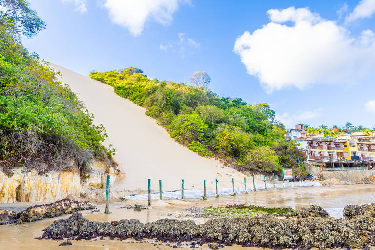 morro deslizado de areia, ladeado por vegetação com sobrados ao fundo e mar à frente