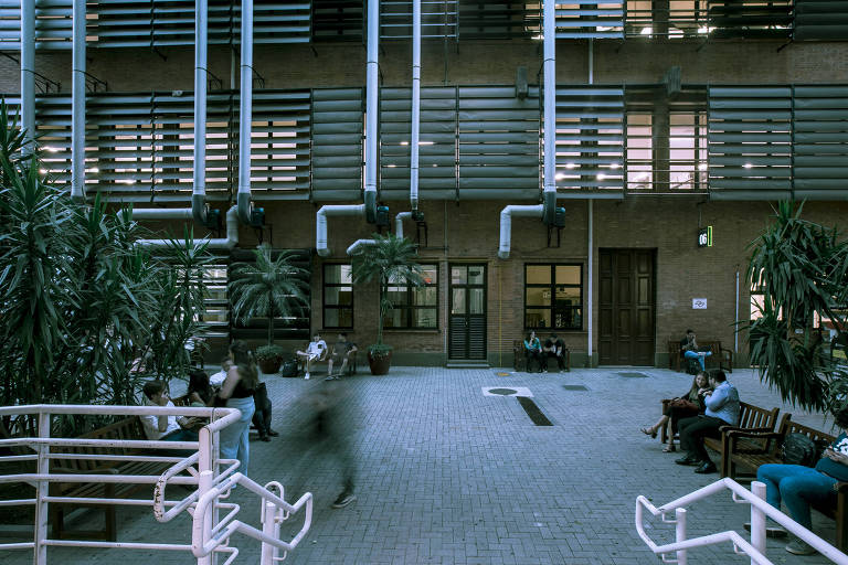 A imagem mostra o interior de um edifício, parte de uma universidade. O ambiente é amplo, com piso claro e algumas pessoas sentadas em bancos distribuídos pelo espaço. Nas paredes, há janelas cobertas por estruturas de ripas horizontais. Há plantas em em vasos.