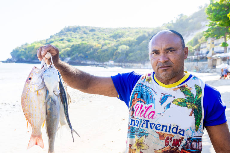 homem segura peixes em praia, com morro ao fundo