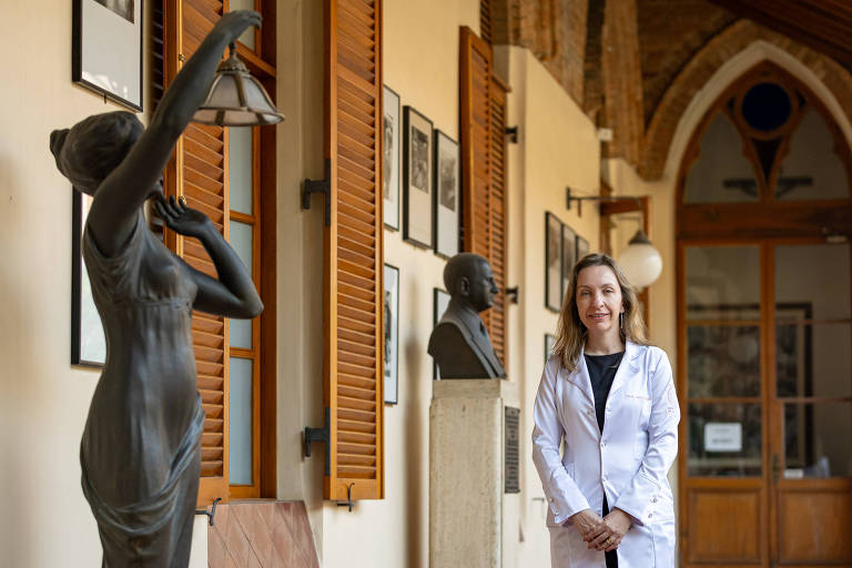 A imagem mostra uma mulher sorridente vestindo um jaleco branco. Ela está em um ambiente com uma arquitetura clássica: ao fundo, há portas e janelas com molduras de madeira e arcos em estilo tradicional. Também são visíveis algumas esculturas: uma figura humana estilizada e um busto.