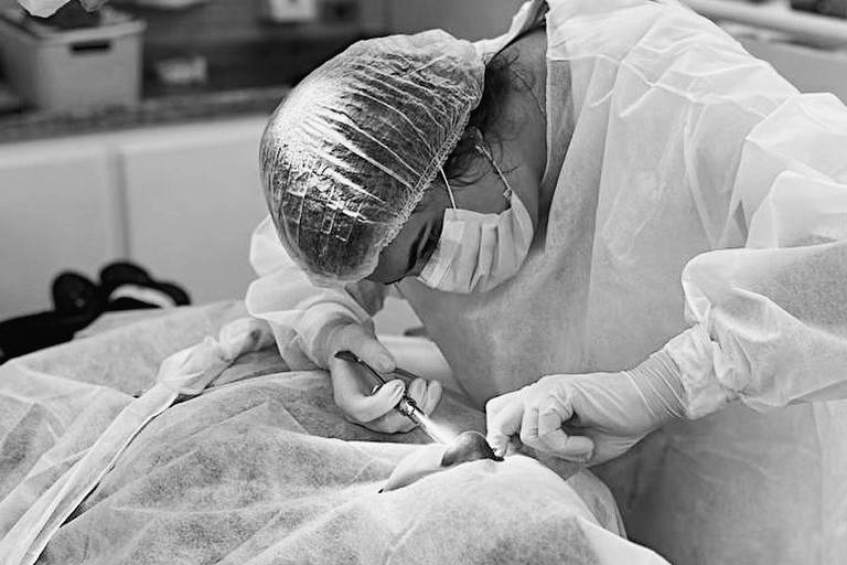 A imagem mostra um profissional de saúde realizando um procedimento cirúrgico. A pessoa está vestida com um avental cirúrgico, luvas, máscara e touca. Ela está concentrada em uma área coberta por um lençol cirúrgico, utilizando um instrumento cirúrgico.