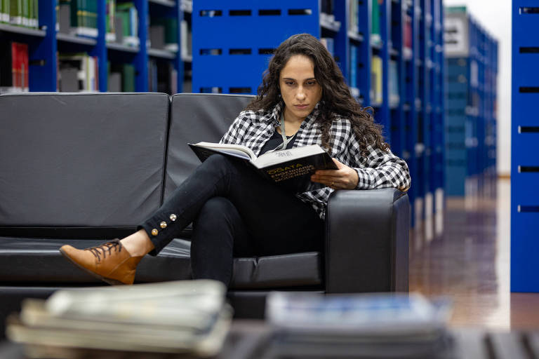 Uma mulher sentada em um sofá na biblioteca, lendo um livro. Ela está vestindo uma camisa xadrez e calças pretas, com sapatos marrons. Ao fundo, prateleiras azuis cheias de livros. Na frente, há alguns livros empilhados.