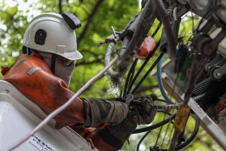 Um trabalhador está em uma plataforma elevada， usando um capacete branco e roupas de proteção laranja. Ele está manipulando fios e cabos， com árvores verdes ao fundo， sugerindo que a manutenção está sendo realizada em uma área externa.