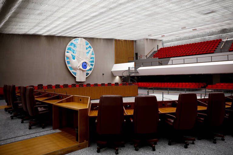 Uma grande sala com uma mesa circular de madeira, com poltronas iguais e vazias lado a lado e um auditório acima, com poltronas vermelhas. Na parede lateral, um quadro redondo com a escultura de uma mão e um olho azul ao meio.