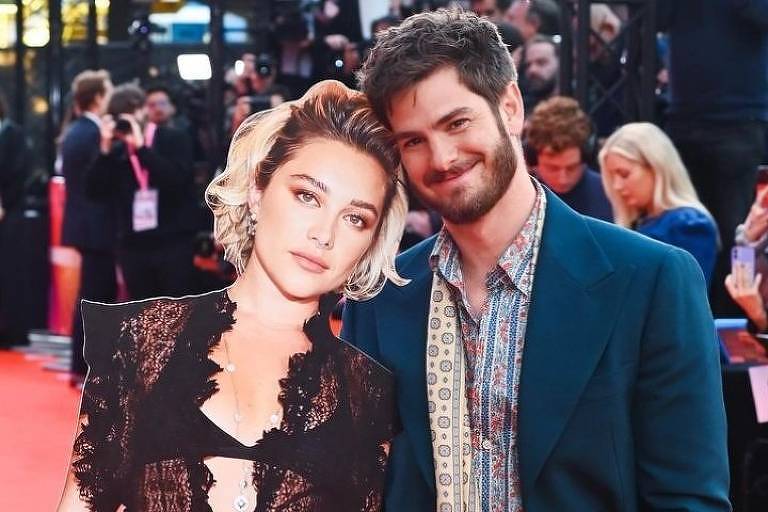 A imagem mostra um homem posando ao lado de um recorte de papel de uma mulher em um evento de tapete vermelho. O homem está sorrindo e vestindo um terno azul escuro com uma camisa estampada. O recorte da mulher usa um vestido preto com detalhes em renda e um colar. O fundo é composto por pessoas e câmeras, indicando um evento de gala.