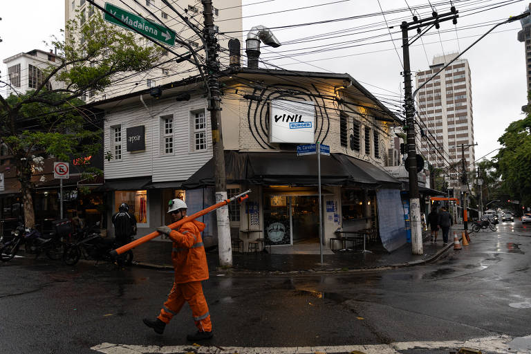 A imagem mostra um cruzamento em uma cidade durante um dia chuvoso. Um homem vestido com um uniforme laranja está caminhando com uma vara laranja na mão. Ao fundo, há um prédio de estilo antigo e outros edifícios modernos. O chão está molhado devido à chuva, e há fios elétricos visíveis acima. A sinalização de trânsito e uma parada de ônibus também estão presentes.