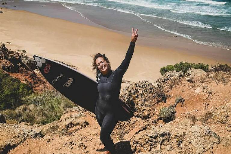 Uma surfista está em uma rocha, sorrindo e fazendo um sinal de paz com a mão direita. Ela usa um traje de neoprene preto e segura uma prancha de surf preta com detalhes brancos. Ao fundo, vê-se uma praia de areia clara e ondas do mar, com um céu azul e algumas nuvens. A vegetação ao redor é baixa e rochosa.