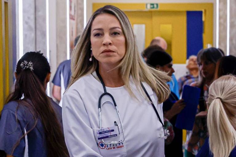 A imagem mostra uma médica com cabelo longo e liso, usando um jaleco branco e um estetoscópio ao redor do pescoço. Ela está caminhando em um corredor de hospital, com um olhar sério. Ao fundo, há várias pessoas, incluindo outros profissionais de saúde e pacientes. As paredes do corredor são de cores claras, e há uma porta amarela ao fundo.