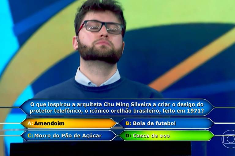 A imagem mostra um homem com barba e óculos, em um cenário de quiz. Ele parece pensativo, com a cabeça levemente inclinada para cima. Ao fundo, há um design colorido. Na parte inferior da imagem, há uma pergunta sobre o que inspirou a arquiteta Chu Ming Silveira a criar o design do protetor telefônico, com quatro opções de resposta: A) Amendoim, B) Bola de futebol, C) Morro do Pão de Açúcar, D) Casca de ovo.