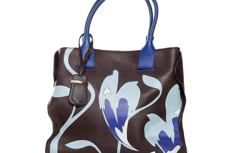 A imagem mostra uma bolsa de couro marrom com alças azuis. A bolsa apresenta uma estampa floral em tons de azul claro e azul escuro, com folhas e flores estilizadas. O logotipo 'TOMS' está visível na parte frontal da bolsa, e há um pequeno detalhe em couro na alça.