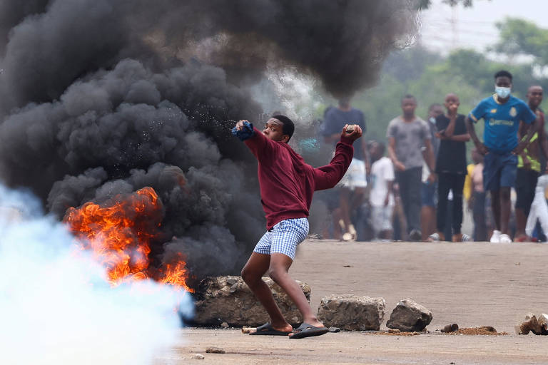 A imagem mostra um jovem em uma rua durante um protesto, lançando um objeto em direção a uma fogueira. Ao fundo, há uma nuvem de fumaça preta e várias pessoas observando a cena. O jovem está vestido com um moletom vermelho e shorts, enquanto a fogueira emite chamas laranjas. O ambiente parece tenso, com a presença de fumaça e destroços no chão.