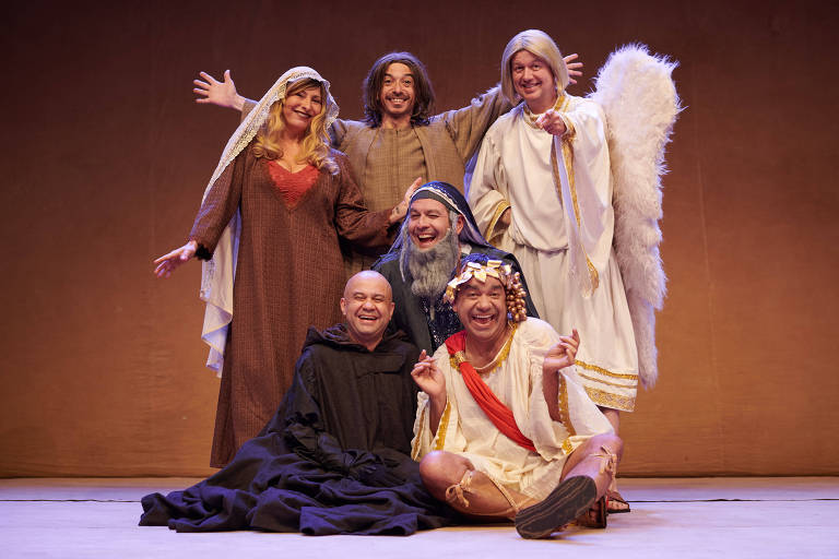 Um grupo de seis pessoas posa para a foto no palco de um teatro. Eles estão vestidos com trajes variados， incluindo uma mulher com cabelo loiro e vestido longo， um homem com uma túnica e barba， um anjo com asas， e outros personagens. O fundo é de cor marrom claro.
