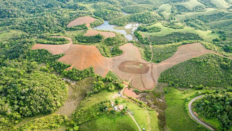 Imagem aérea de uma área rural com vegetação densa e campos desmatados. A paisagem apresenta uma mistura de áreas verdes e terrenos arados, com um pequeno lago visível no centro. Estradas de terra serpenteiam pela região, e há uma construção próxima a um corpo d'água. O solo arado é de cor marrom claro, contrastando com o verde das árvores ao redor.