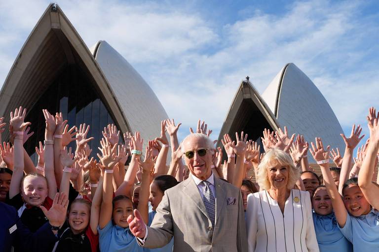 A imagem mostra um grupo de crianças levantando as mãos em frente à Ópera de Sydney. No centro, um homem de terno claro e óculos escuros, ao lado de uma mulher com cabelo loiro e blusa listrada. Ao fundo, as icônicas estruturas da Ópera de Sydney sob um céu azul com algumas nuvens.