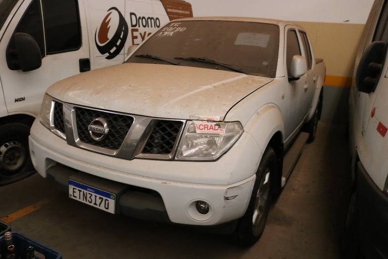 A imagem mostra uma caminhonete da marca Nissan, de cor branca, estacionada em um local com outros veículos ao fundo. O veículo apresenta um design robusto, com uma grade frontal proeminente e faróis grandes. A caminhonete parece estar em um estado usado, com algumas marcas visíveis na pintura.