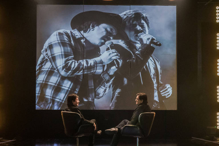 A imagem mostra um estúdio de gravação com duas pessoas sentadas em cadeiras giratórias, conversando. Ao fundo, uma tela exibe uma foto em preto e branco de dois cantores se apresentando. O ambiente é iluminado por luzes brilhantes dispostas nas laterais e no teto. Um cameraman está visível à direita, operando uma câmera.