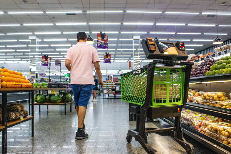 homem de costas caminha em corredor de frutas de supermercado com um carrinho de compras verde à frente. o carrinho carreta maquinas de cartão de crédito