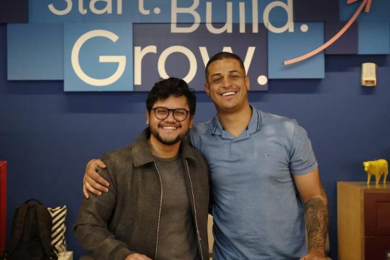 Na imagem, dois homens estão sorrindo e posando juntos em um ambiente de trabalho. O fundo apresenta uma parede azul com as palavras 'Start. Build. Grow.' em letras brancas. O homem à esquerda usa óculos e uma jaqueta escura, enquanto o homem à direita está vestido com uma camiseta azul. Ambos estão em frente a uma mesa de madeira.