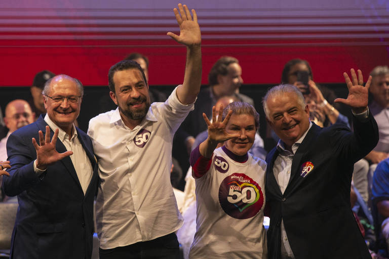 Quatro pessoas estão em um evento político， sorrindo e acenando com a mão levantada. Eles estão vestidos de forma casual， com uma pessoa usando uma camiseta branca com o número 50. O fundo é decorado com cores vibrantes， e há uma plateia visível ao fundo.