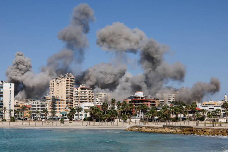 A imagem mostra uma área urbana à beira-mar com prédios e uma praia. No fundo, há grandes nuvens de fumaça cinza subindo, indicando uma possível explosão ou incêndio. O céu está claro e azul, contrastando com a fumaça. A vegetação é visível na área costeira.