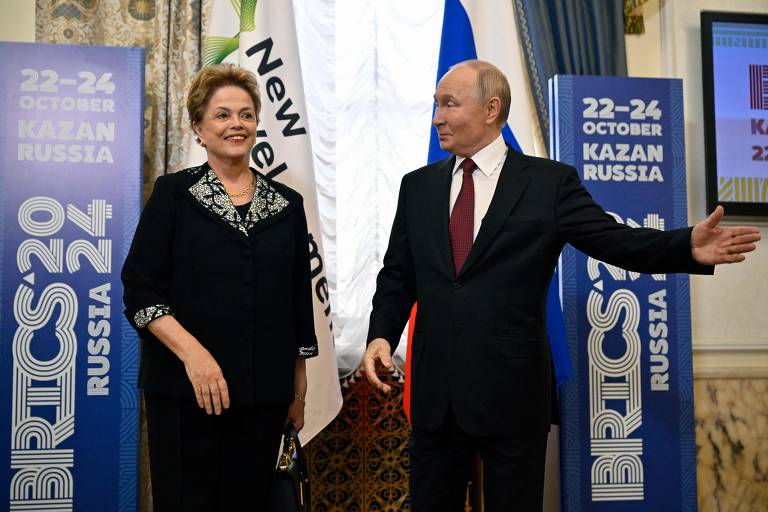 A imagem mostra duas pessoas em um evento relacionado ao BRICS 2024. À esquerda, uma mulher com cabelo cacheado e vestido escuro, e à direita, um homem com cabelo curto e terno escuro. Eles estão em frente a banners que indicam o evento, com as datas de 22 a 24 de outubro em Kazan, Rússia. O fundo é decorado com cortinas e bandeiras.