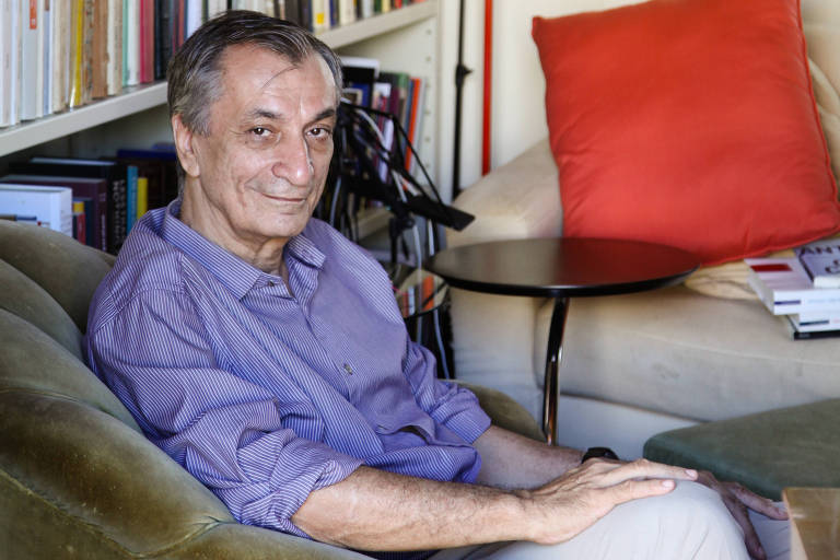 Um homem está sentado em um sofá verde claro em uma sala com estantes cheias de livros. Ele usa uma camisa roxa e tem cabelo grisalho