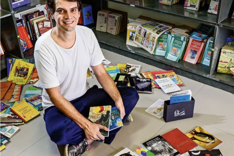 A imagem mostra um homem de camiseta branca e calça azul sentado no chão, cercado por vários livros infantis e juvenis. Ele está segurando dois livros nas mãos, enquanto outros estão espalhados ao seu redor e em uma estante ao fundo. O cenário parece ser de uma biblioteca ou espaço dedicado à leitura, com livros dispostos de forma casual. O homem sorri para a câmera, criando uma atmosfera amigável e acolhedora.