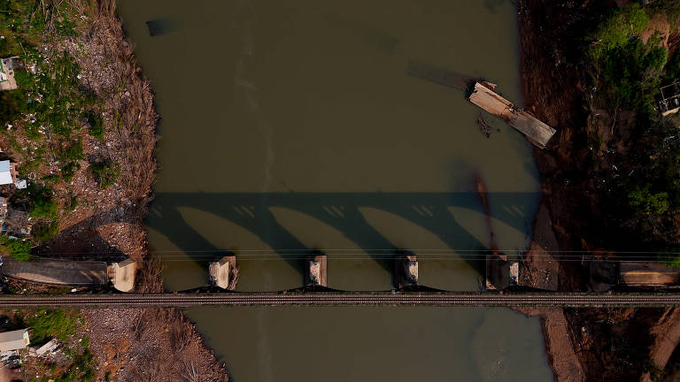 A imagem mostra uma vista aérea de uma ponte de ferro que atravessa um rio. A ponte possui várias vigas de suporte visíveis e projeta sombras sobre a água. À esquerda, há uma área com vegetação e, à direita, uma margem com terra exposta e detritos. O rio apresenta uma coloração esverdeada e está parcialmente obstruído por um barco encalhado.