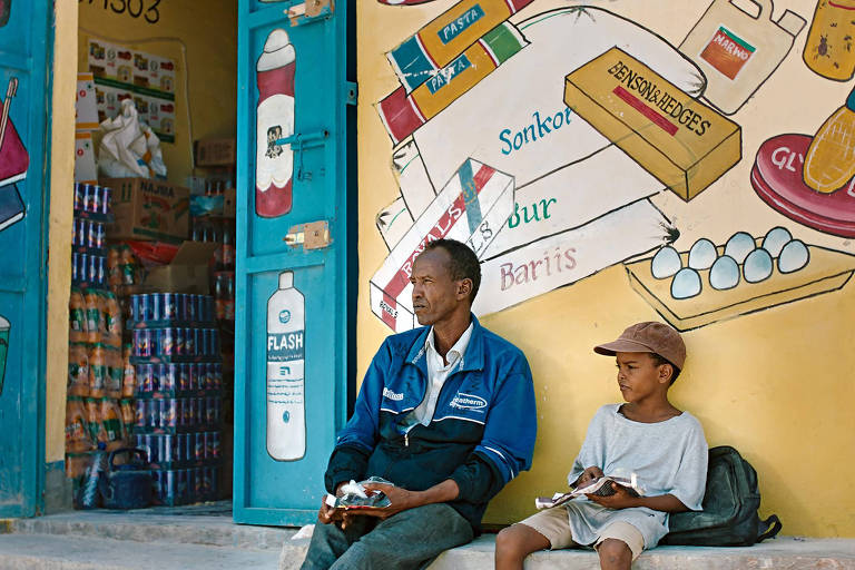 A imagem mostra um homem e uma criança sentados em frente a uma loja. O homem, vestido com uma jaqueta azul e calças, observa pensativo. A criança, usando uma camiseta cinza e um boné, segura algo nas mãos. Ao fundo, a loja tem portas azuis abertas, revelando prateleiras com produtos e uma parede decorada com ilustrações de itens como livros, alimentos e bebidas.