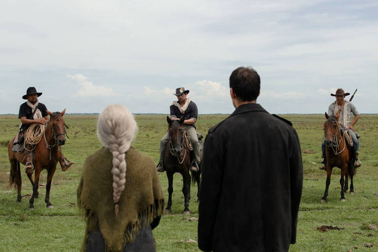 A imagem mostra um confronto no campo aberto, onde quatro homens a cavalo estão em posição de destaque. Dois deles estão à esquerda, montados em cavalos marrons, enquanto os outros dois estão à direita, um deles segurando uma arma. No centro, uma mulher de cabelo longo e prateado, vestindo um xale verde, observa a cena. Um homem de costas, usando um casaco preto, também está presente, voltado para os cavaleiros. O céu está parcialmente nublado e o campo é vasto e verde.