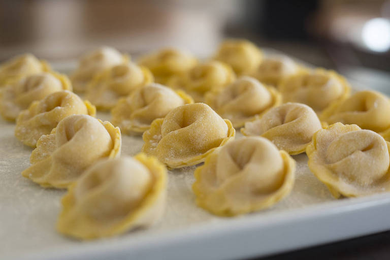 A imagem mostra uma bandeja com várias porções de massa de ravioli, dispostas em fileiras. As massas têm uma forma arredondada e estão levemente polvilhadas com farinha, apresentando uma cor amarelada. O fundo da imagem é desfocado, destacando as massas em primeiro plano.