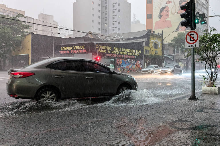 A imagem mostra um carro cinza atravessando uma rua alagada durante uma forte chuva. A água está acumulada na pista, e o veículo está parcialmente submerso. Ao fundo, há prédios altos e um letreiro visível, além de semáforos e placas de trânsito. A cena é marcada por um clima de tempestade, com nuvens escuras.