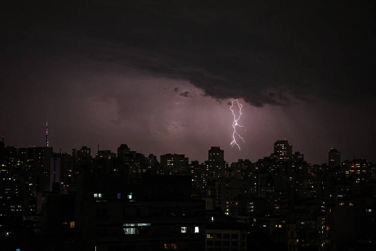 A imagem mostra uma cidade à noite, com muitos prédios iluminados. No céu, nuvens escuras estão presentes, e um raio é visível descendo em direção ao solo. A iluminação da cidade contrasta com a escuridão do céu.