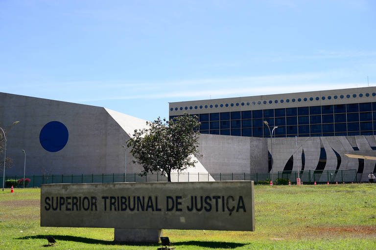 Imagem do Superior Tribunal de Justiça, um edifício moderno com uma estrutura arquitetônica distinta. À frente, há uma placa com o nome da instituição, e um pequeno arbusto está presente no gramado. O céu está claro e azul, e o edifício apresenta formas geométricas e janelas grandes.