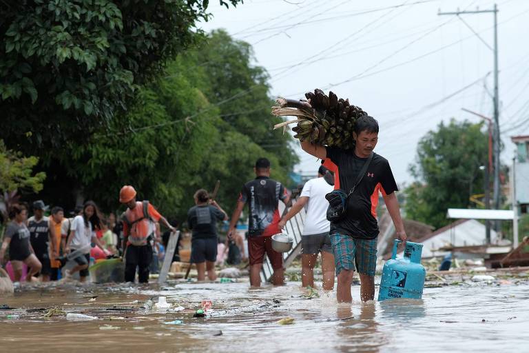 A imagem mostra uma cena de inundação em uma área urbana, com várias pessoas caminhando em águas alagadas. Um homem carrega um feixe de bananas sobre os ombros, enquanto outros indivíduos estão ao fundo, alguns com objetos nas mãos. A vegetação ao redor é densa e há postes de eletricidade visíveis. O céu está nublado, indicando um clima chuvoso.