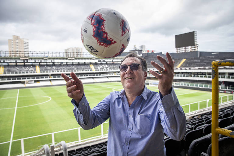 Um homem sorridente está em um estádio de futebol, jogando uma bola para cima. Ele usa óculos escuros e uma camisa de botão azul. Ao fundo, vê-se o campo de futebol verde e as arquibancadas do estádio, sob um céu nublado.