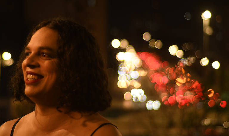 Mulher de pele branca e cabelo preto cacheado sorri em ambiente aberto à noite com luzes de faróis desbotados ao fundo