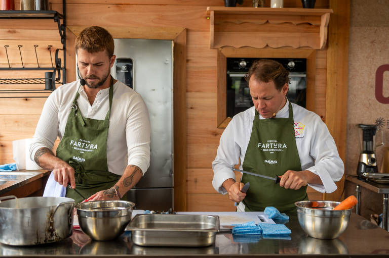 Foto mostra dois homens em uma cozinha, ambos usando aventais verdes. O homem branco à esquerda tem cabelo curto e barba, e está sorrindo enquanto segura um pano de prato. O homem branco à direita, com cabelo loiro e ondulado, também sorri. Ao fundo, há um fogão e uma geladeira prateada. Na mesa, há utensílios de cozinha, incluindo tigelas de metal e um recipiente com cenouras.