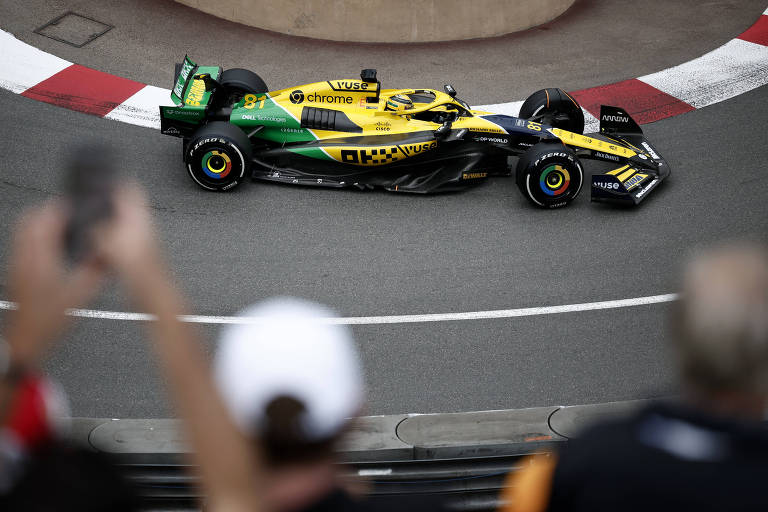 Um carro de Fórmula 1, predominantemente nas cores amarelo e verde, está fazendo uma curva em uma pista de corrida em Mônaco. Ao fundo, é possível ver pessoas assistindo à corrida, algumas com os braços levantados, segurando celulares.