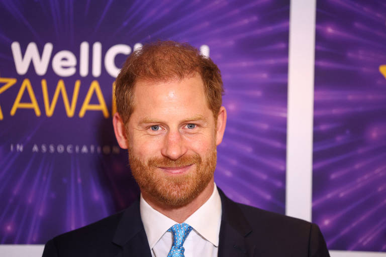 Harry, um homem branco com cabelo ruivo e barba, sorrio. Ele usa um terno escuro e uma gravata azul clara. O fundo é decorado com um padrão roxo e contém o texto 'WellChild Awards'.