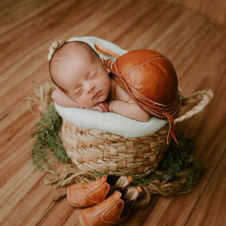 Um bebê recém-nascido dormindo em uma cesta de vime. O bebê está deitado sobre um tecido branco, com um chapéu de couro marrom ao lado. Ao fundo, há um piso de madeira e algumas folhas verdes. Perto da cesta, há um par de sapatos de couro marrom.