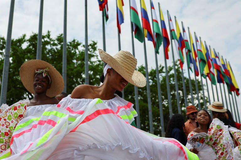 A imagem mostra um grupo de pessoas dançando em trajes tradicionais coloridos, com saias brancas e detalhes em neon. Elas usam chapéus de palha e estão em frente a uma fileira de bandeiras da Colômbia. O ambiente é festivo, com árvores ao fundo e um céu parcialmente nublado.