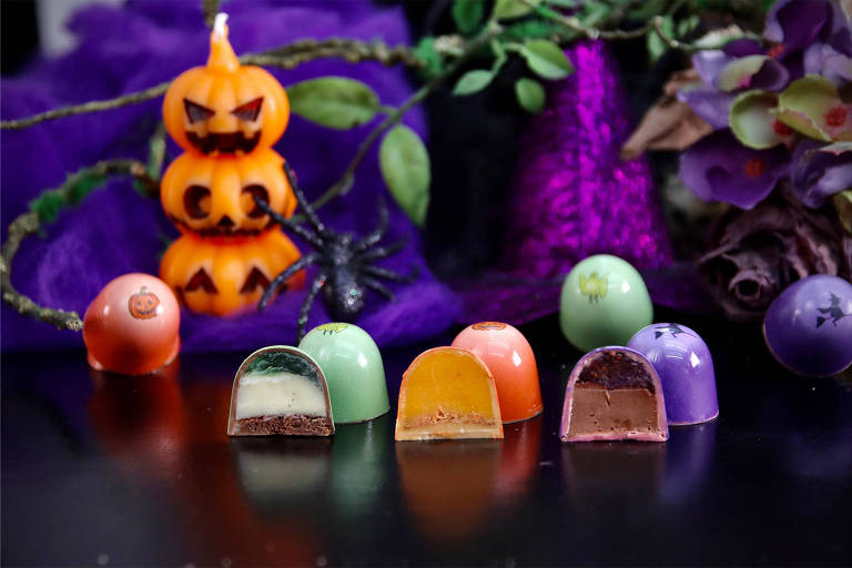 A imagem apresenta uma variedade de chocolates decorados, com um fundo temático de Halloween. Há uma vela em forma de abóbora empilhada, com rostos assustadores, e uma aranha de plástico. Os chocolates têm cores variadas, como verde, laranja, roxo e rosa, alguns com desenhos de morcegos e abóboras. Um dos chocolates está cortado ao meio, mostrando seu recheio colorido.