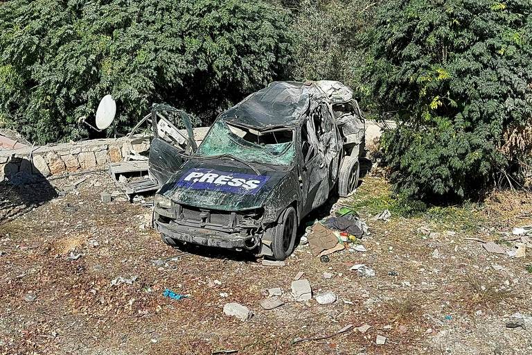 A imagem mostra um veículo danificado, aparentemente abandonado, em um terreno com vegetação ao fundo. O carro é de cor escura e possui uma inscrição 'PRESS' em destaque na lateral. O veículo está em estado de deterioração, com janelas quebradas e a carroceria amassada. Ao redor, há lixo e detritos espalhados pelo chão.