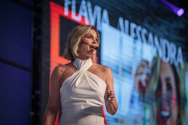 Uma mulher com cabelo curto e loiro， vestindo um vestido branco， está em um palco durante uma apresentação. Ela está segurando um microfone e parece estar falando para a audiência. Ao fundo， há um telão com o nome 039;FLÁVIA ALESSANDRA039; e imagens coloridas.