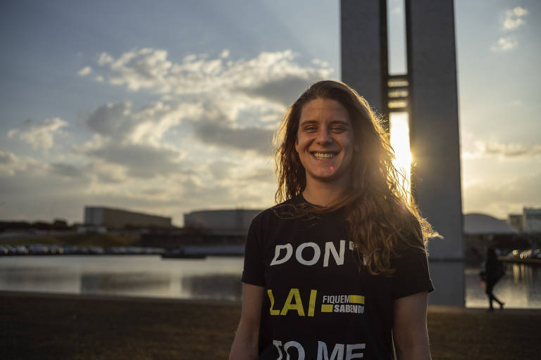 A imagem mostra uma mulher sorrindo ao ar livre, com o céu ao fundo no momento do pôr do sol. Ela está vestindo uma camiseta preta com a frase "DON'T LAI TO ME", associada à campanha do Fiquem Sabendo sobre transparência e acesso à informação. Ao fundo, é possível ver um monumento icônico de Brasília, sugerindo que a foto foi tirada na capital federal. A iluminação do sol cria um efeito de destaque, iluminando os cabelos e parte do rosto da mulher, conferindo um tom caloroso e vibrante à foto.