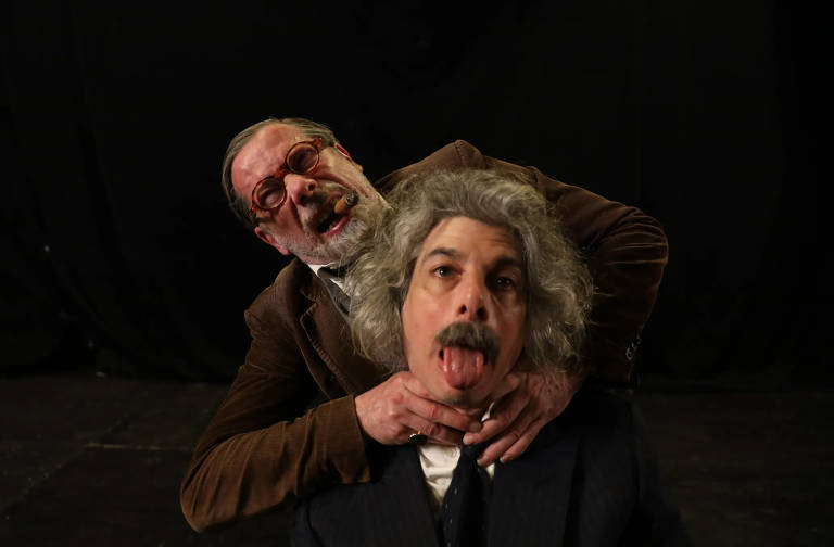 A imagem mostra dois homens em uma cena teatral. Um homem, com óculos e barba, está atrás do outro, segurando seu pescoço. O homem da frente tem cabelo grisalho e está fazendo uma expressão exagerada, com a língua para fora. O fundo é escuro, sugerindo um ambiente de palco.