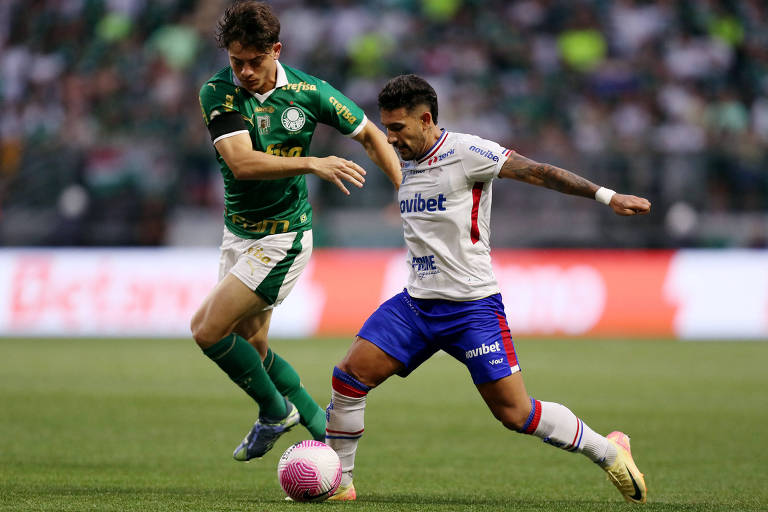 Dois jogadores de futebol estão em campo durante uma partida. O jogador à esquerda veste uma camisa verde e branca, do Palmeiras, enquanto o jogador à direita usa uma camisa branca com detalhes em azul e vermelho, do Fortaleza. O jogador da esquerda tenta desarmar o jogador da direita