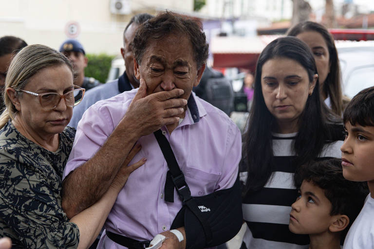 A imagem mostra um grupo de pessoas em um momento emocional. Um homem de camisa rosa， com um braço imobilizado， está visivelmente emocionado， cobrindo o rosto com a mão. Ao seu redor， há uma mulher mais velha， uma mulher com cabelo escuro e liso， e duas crianças， uma delas com cabelo curto e a outra com cabelo mais longo. O ambiente parece ser ao ar livre， com outras pessoas ao fundo.