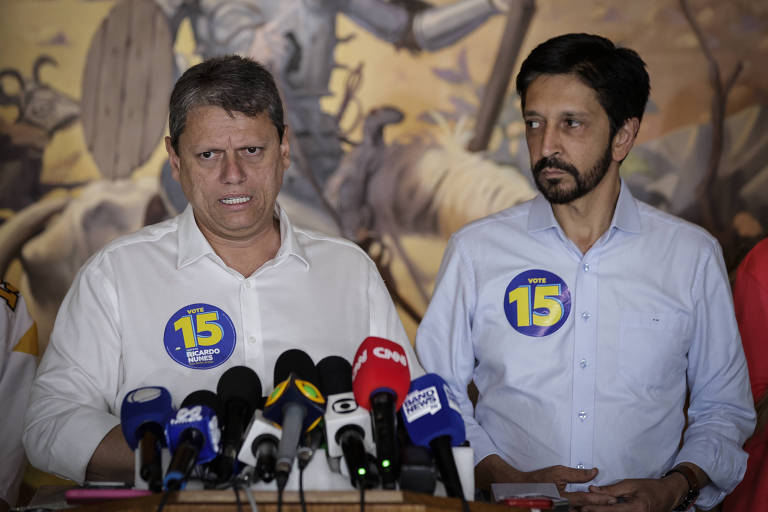 Dois homens estão em uma coletiva de imprensa, um deles está à esquerda, vestindo uma camisa branca e o outro à direita, com uma camisa azul clara. Ambos têm adesivos com o número 15 em seus peitos. Há vários microfones na mesa à frente deles e um mural ao fundo com uma imagem de uma cena histórica.